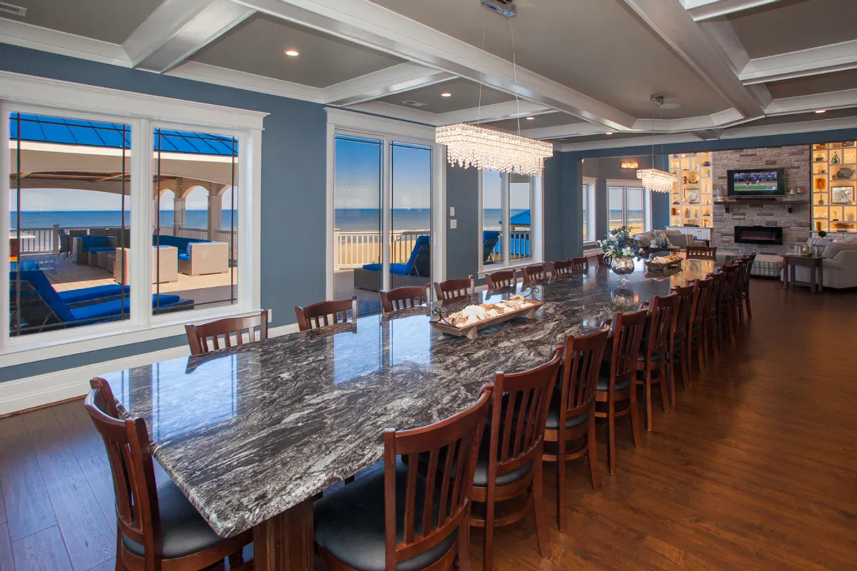 A spacious dining area with a large table sits beside expansive windows revealing an ocean view. A chandelier and fireplace add warmth and elegance, making it an ideal spot for shared meals, celebrations, or casual gatherings.