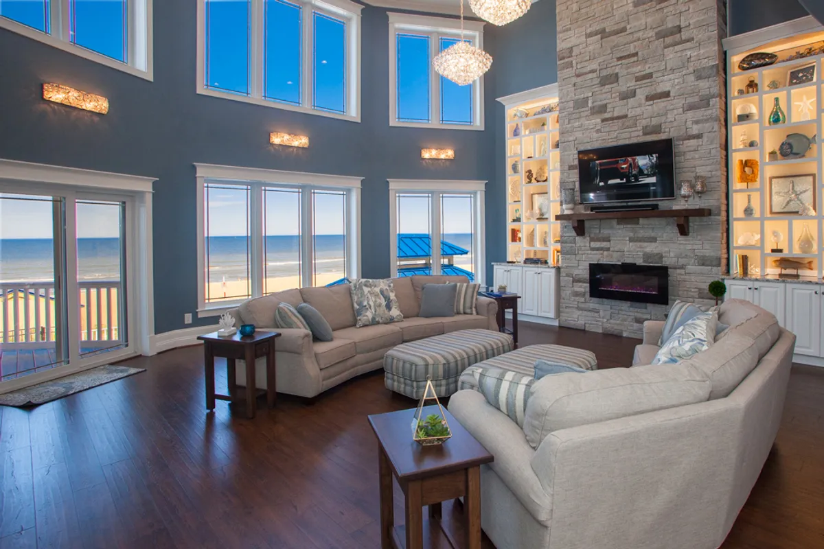 This interior shot features expansive windows with stunning beach views, a luxurious stone fireplace, chandeliers, and comfortable seating arranged for relaxation or family gatherings. The atmosphere is sophisticated yet cozy, and while the kitchen is not directly visible, the sense of an inviting, interconnected interior environment is palpable.