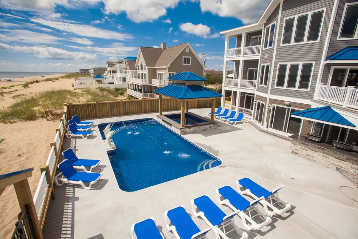 A bright, cheerful pool area featuring lounge chairs, a gazebo, and direct beach access. The space is designed for sunbathing, outdoor dining, and family gatherings, set amidst beautifully maintained grounds with ocean vistas in the background.