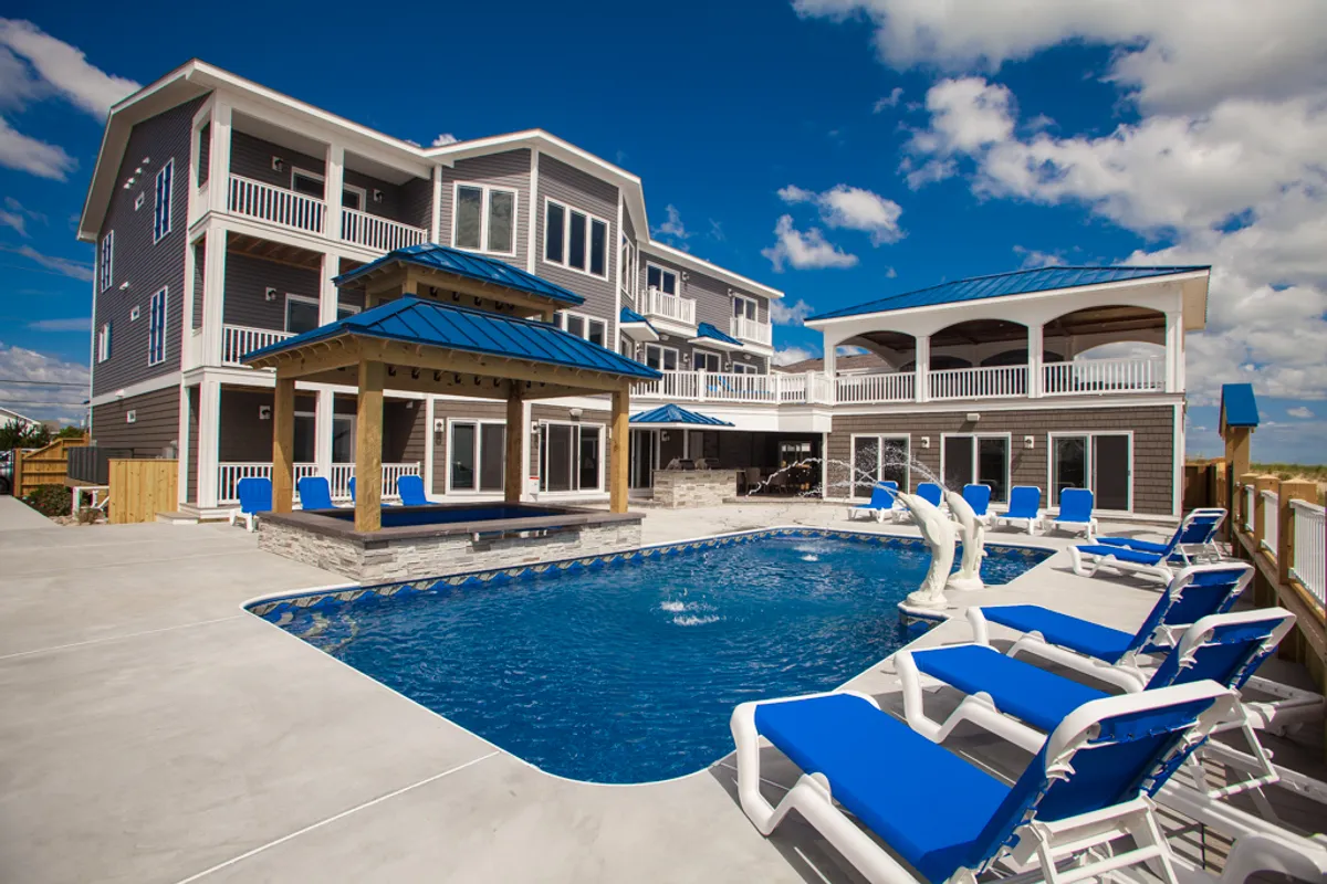 The image features the inviting exterior of a modern three-story condo complex in Mesa, Arizona, centered around a sparkling swimming pool. Comfortable outdoor seating under a covered pergola invites relaxation, while balconies offer sunset views. The scene radiates a tranquil, resort-like atmosphere perfect for families or groups.